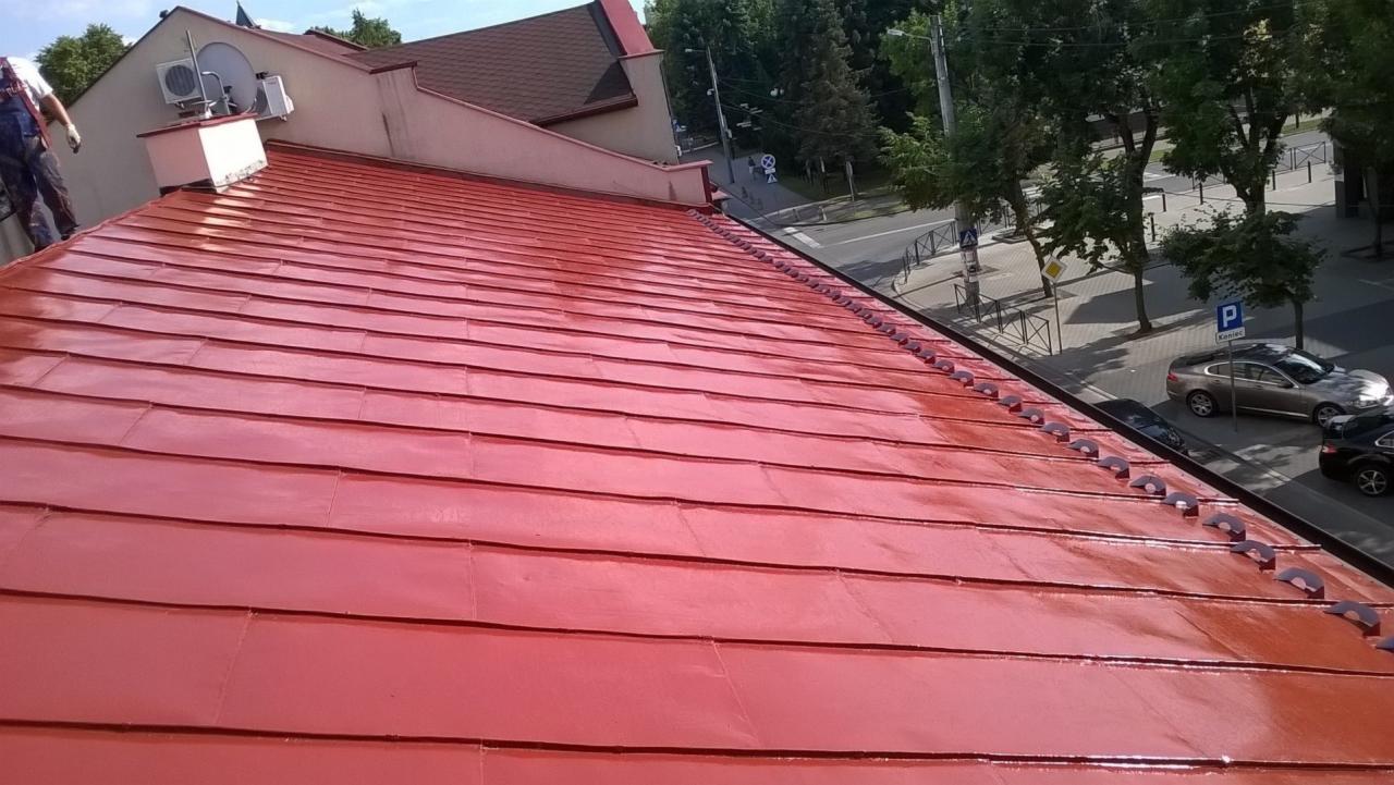 Czerwony, błyszczący dach z blachy dachówkowej po czyszczeniu, widok z góry. W tle budynki, drzewa i ulica z zaparkowanymi samochodami, słoneczny dzień.