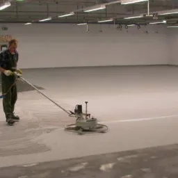 Pracownik w hali wyrównuje posadzkę za pomocą specjalistycznej maszyny. Widoczny fragment świeżo wylanej, jasnej posadzki i gładka, równa powierzchnia.