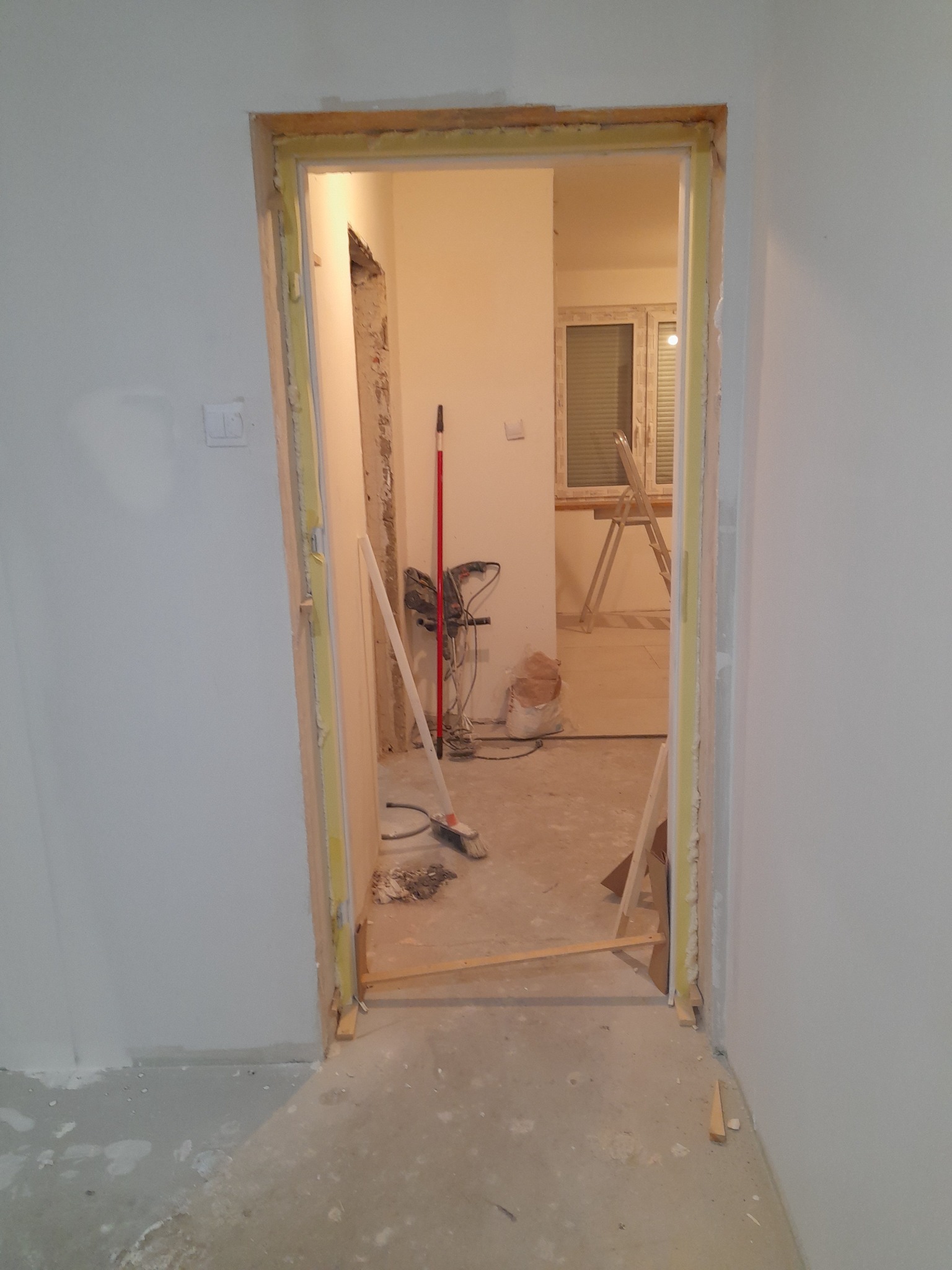 Przygotowanie do montażu ościeżnicy: surowy otwór drzwiowy z drewnianymi klinami i pianką montażową, widoczne narzędzia i drabina w tle.