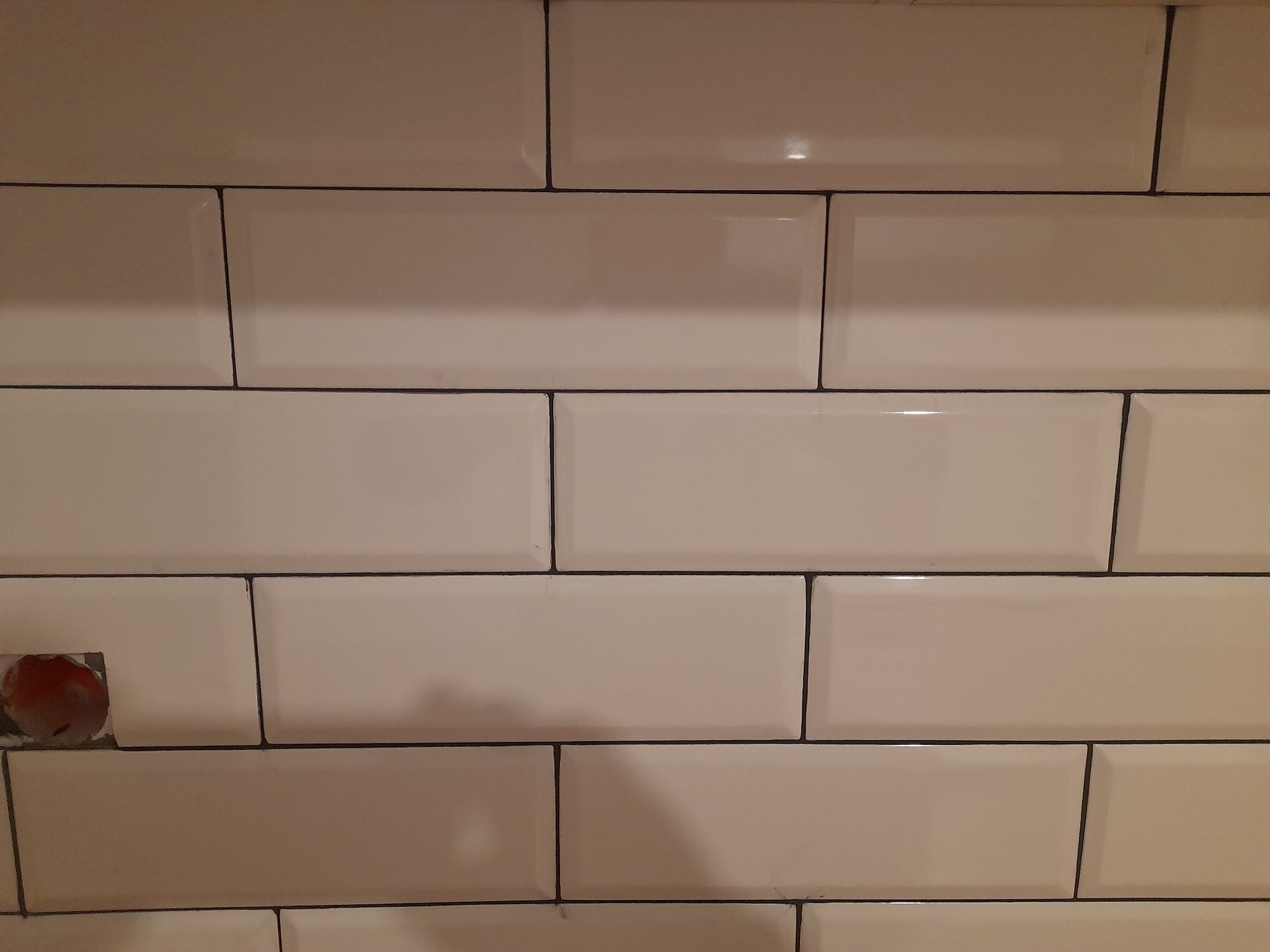 Ułożone, beżowe, prostokątne płytki ceramiczne z ciemną fugą, z widocznym otworem na gniazdko elektryczne w lewym dolnym rogu.