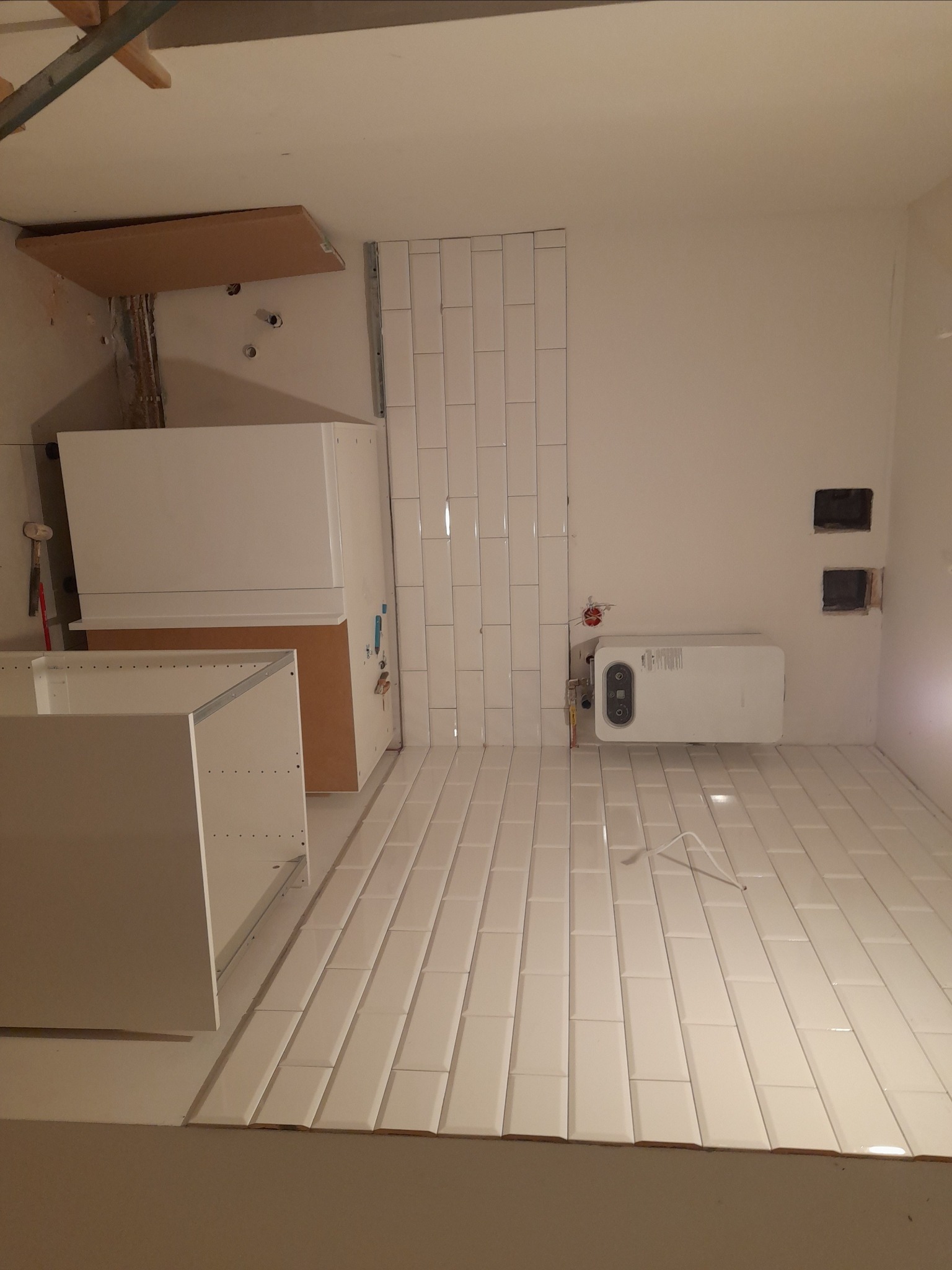 Wnętrze łazienki w trakcie remontu: białe, podłużne płytki ułożone na podłodze i częściowo na ścianie, wiszący biały bojler oraz niezamontowane, białe szafki kuchenne.