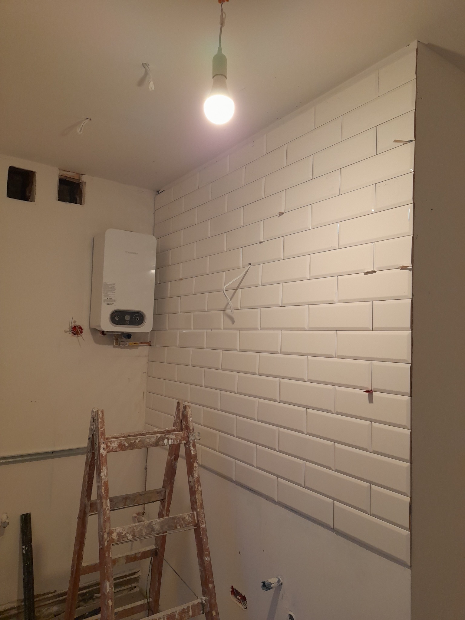 Ściana w trakcie kładzenia białych, podłużnych płytek imitujących cegłę, widoczna drabina malarska, piec gazowy na ścianie i wystające kable elektryczne.
