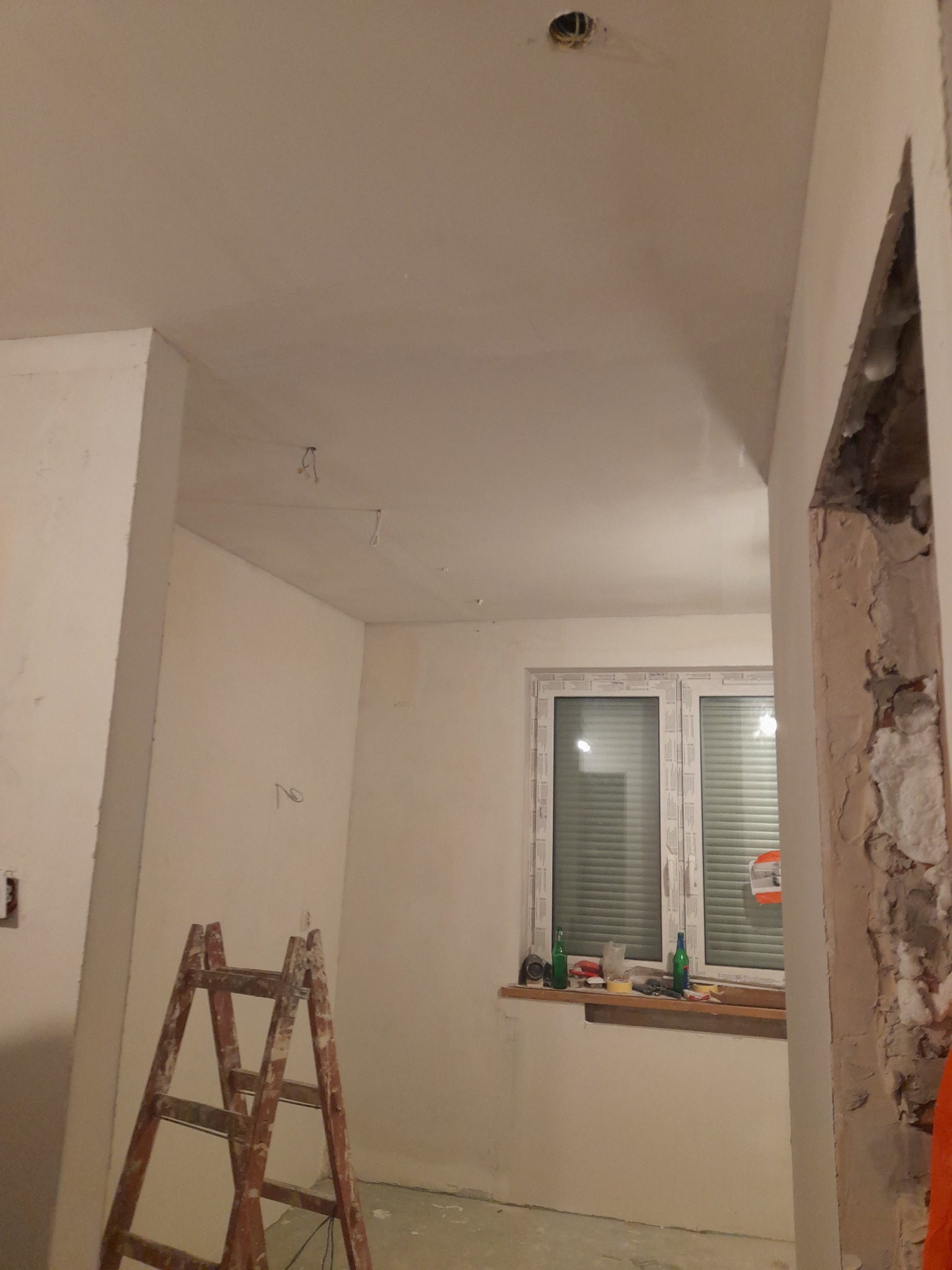 Wnętrze remontowanego pokoju w domu: świeżo otynkowane ściany i sufit z wystającymi kablami, drewniana drabina oparta o ścianę, okno z roletami i butelkami na parapecie, oraz fragment ściany...