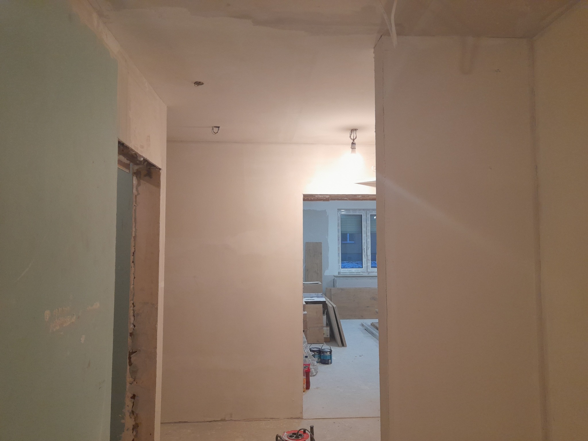 Wnętrze mieszkania w trakcie remontu: surowe ściany, widoczne ślady po usuniętych futrynach, niezagruntowane ściany, materiały budowlane w tle, surowe oświetlenie.
