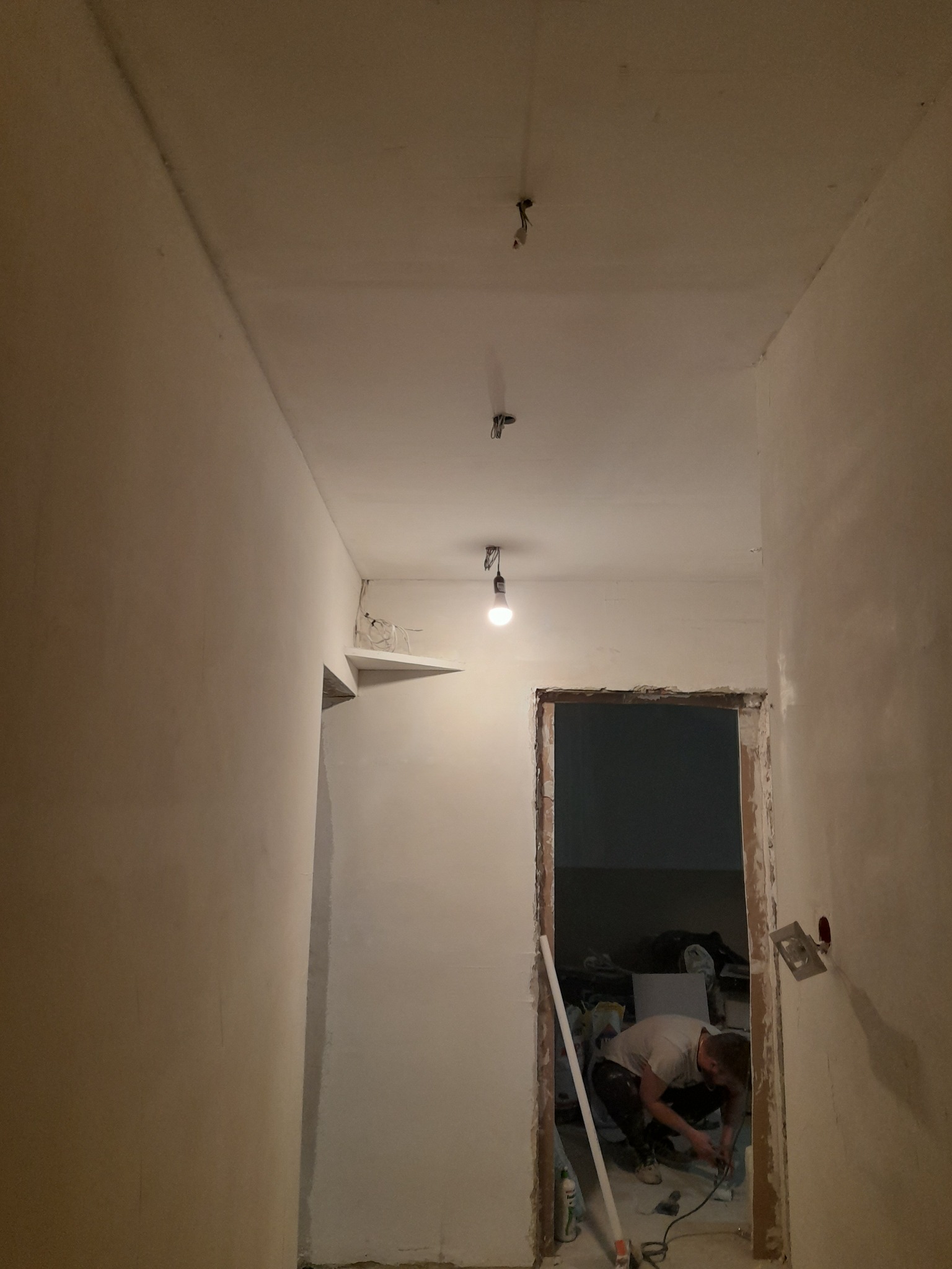 Wnętrze w trakcie szpachlowania ścian, widoczny pracownik z narzędziami, surowy stan wykończenia pod oświetleniem żarówki.