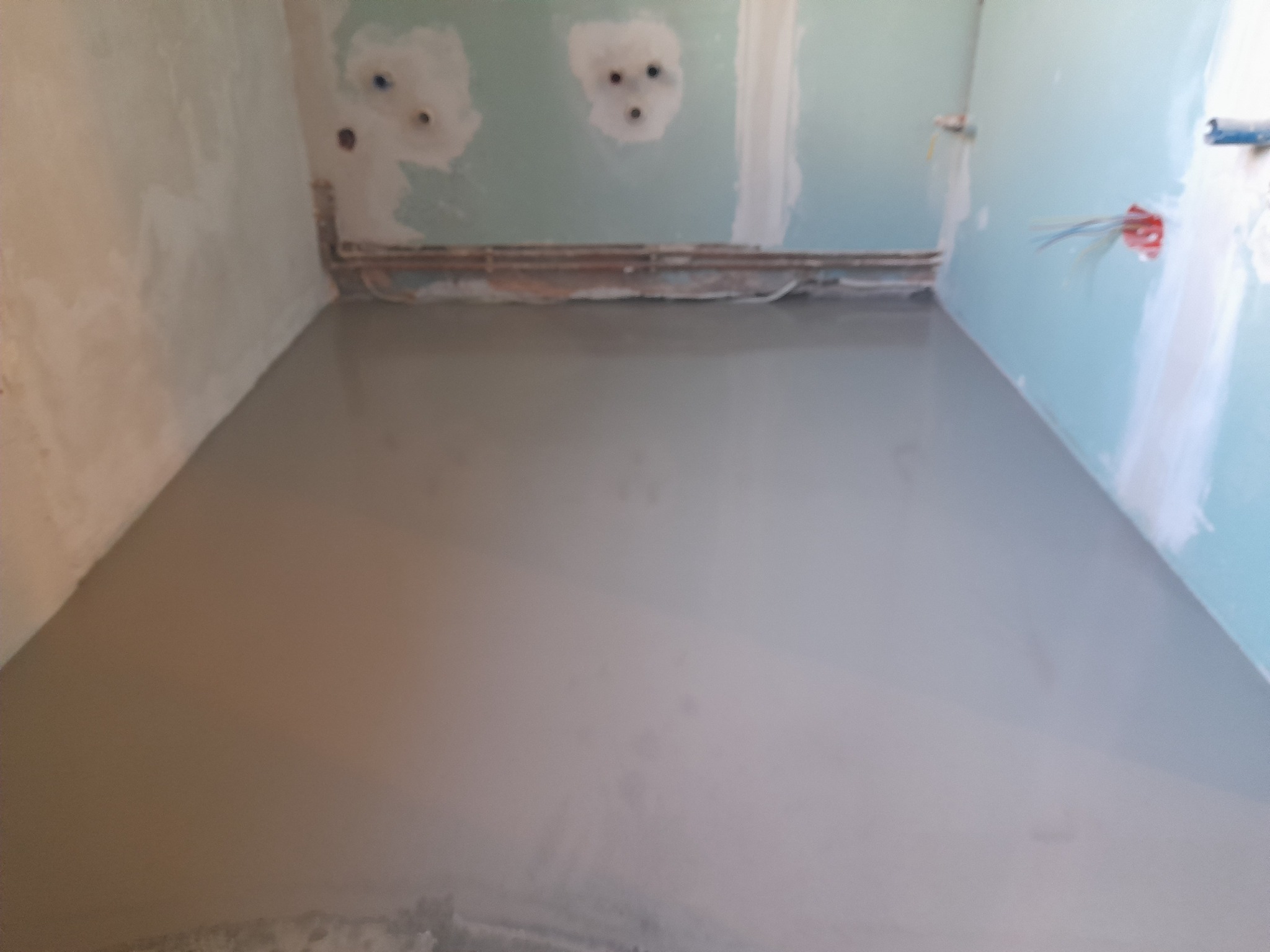 Świeżo wylana, gładka wylewka samopoziomująca w pomieszczeniu z przygotowanymi instalacjami elektrycznymi i rurami grzewczymi na ścianach.