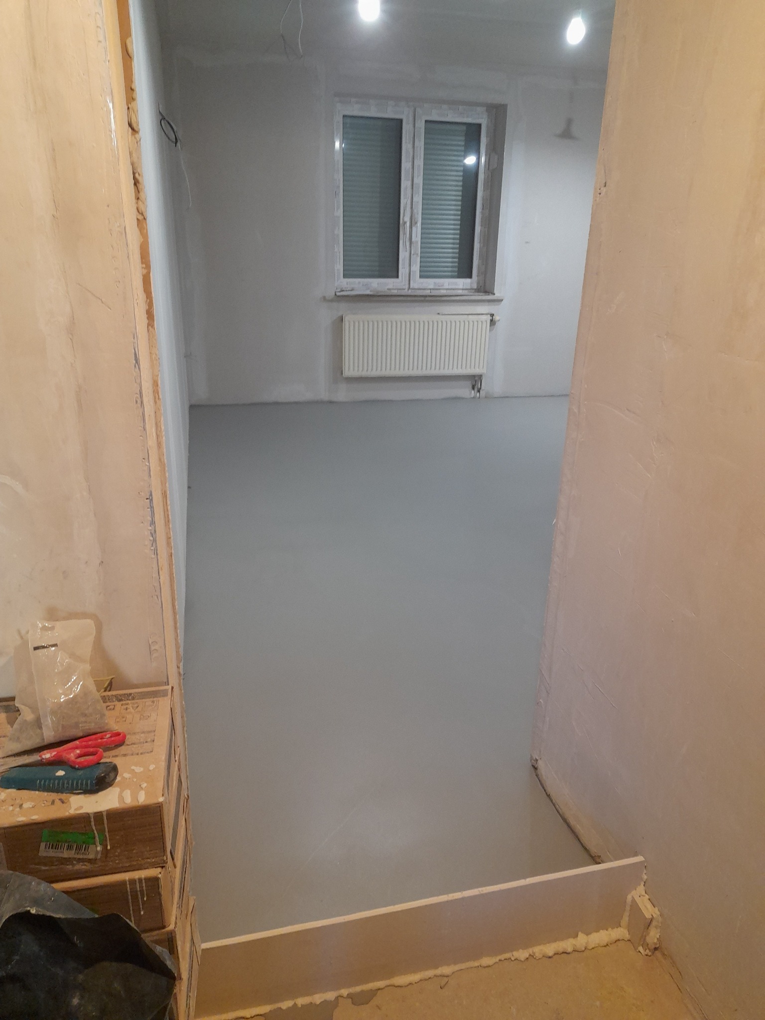 Świeżo wylana, szara wylewka samopoziomująca w pokoju, widoczne okno z grzejnikiem, narzędzia i materiały budowlane w kadrze.