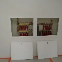 Dwa kolektory ogrzewania podłogowego w białych wnękach, z widocznymi rurami miedzianymi i czerwonymi pętlami, zakryte białymi klapami rewizyjnymi na tle jasnej ściany.