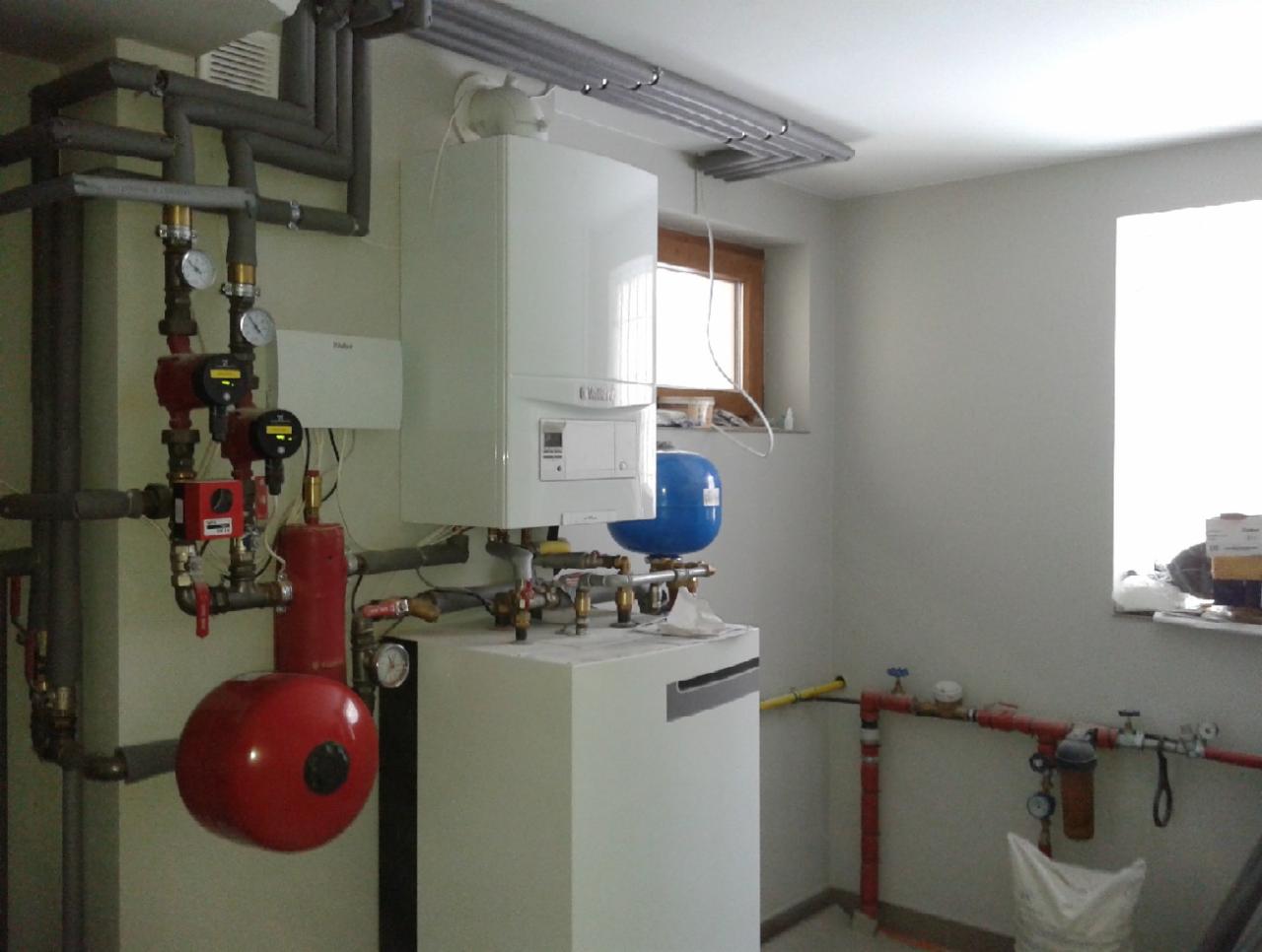 Biały piec gazowy Vaillant z orurowaniem i czerwonym zbiornikiem w kotłowni, widok z boku. Elementy instalacji grzewczej przy ścianie, okno w tle.