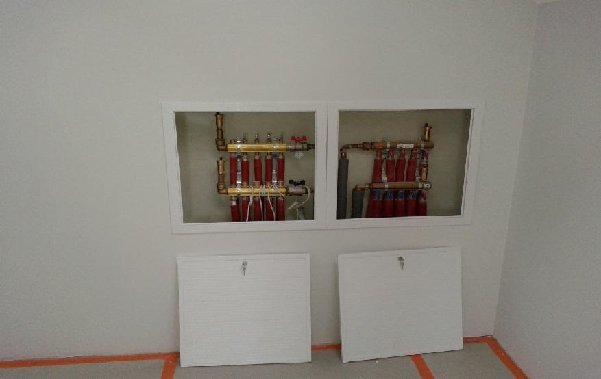 Dwa kolektory ogrzewania podłogowego w białych wnękach, z widocznymi rurami miedzianymi i czerwonymi pętlami, zakryte białymi klapami rewizyjnymi na tle jasnej ściany.