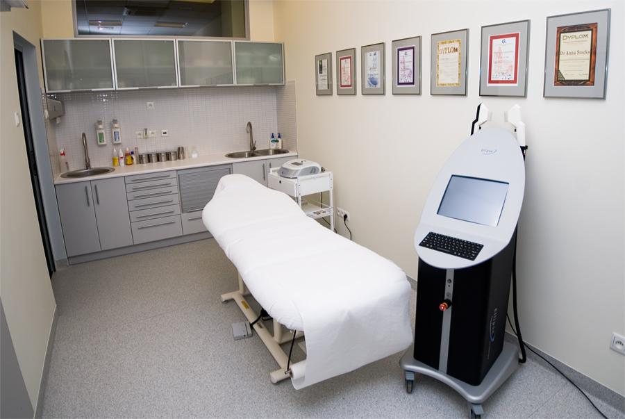 Jasne wnętrze gabinetu medycyny estetycznej z leżanką pokrytą białym prześcieradłem, nowoczesnym urządzeniem do laseroterapii z ekranem dotykowym i klawiaturą, szafkami z umywalkami oraz ramkami...