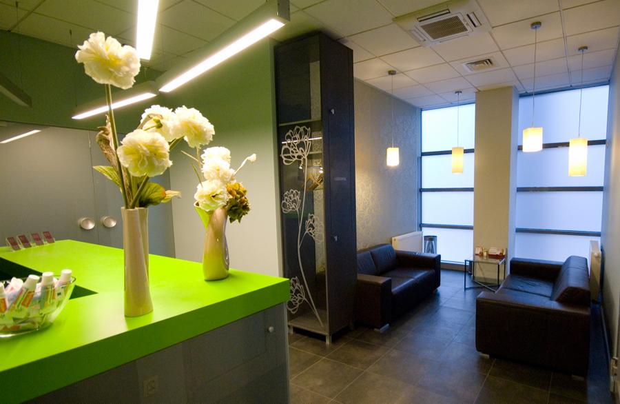 Wnętrze poczekalni prywatnej kliniki z zielonym blatem recepcyjnym, bukietami białych kwiatów w wazonach, ciemnymi sofami i dużymi oknami z żaluzjami.