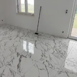 Wnętrze pokoju z nowo położoną, błyszczącą podłogą z płytek imitujących marmur, widoczna miotła oparta o białą ścianę, okno z żaluzjami i wyjście na balkon.