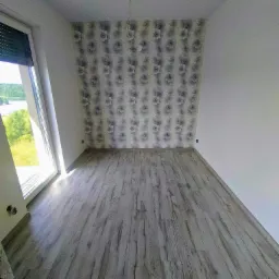 Pusty pokój po remoncie z szarą podłogą panelową imitującą drewno, ściana z tapetą w kwiaty i widok na jezioro przez okno.