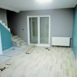 Jasne panele podłogowe ułożone w salonie, widoczne przygotowane schody do wykończenia i ściany pomalowane na szaro i niebiesko, białe okno balkonowe i grzejnik.