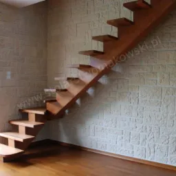 Drewniane schody wspornikowe o ciepłym odcieniu, zamontowane przy ścianie wyłożonej dekoracyjną cegłą w odcieniach beżu, z widocznym adresem strony internetowej na schodach.