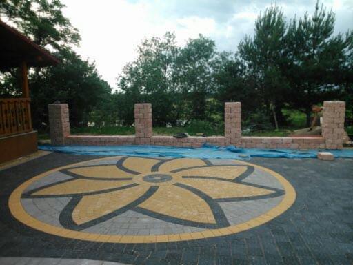 Ozdobny wzór kwiatowy ułożony z kostki brukowej w odcieniach żółci, szarości i czerni na tle częściowo ukończonego muru z cegły, zabezpieczonego folią. W tle widoczny budynek z drewnianą...