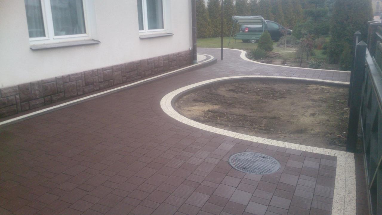 Nowo ułożona, brązowa kostka brukowa z kremowymi obrzeżami tworząca ścieżkę wokół rabaty i wzdłuż elewacji budynku, widoczny właz kanalizacyjny.