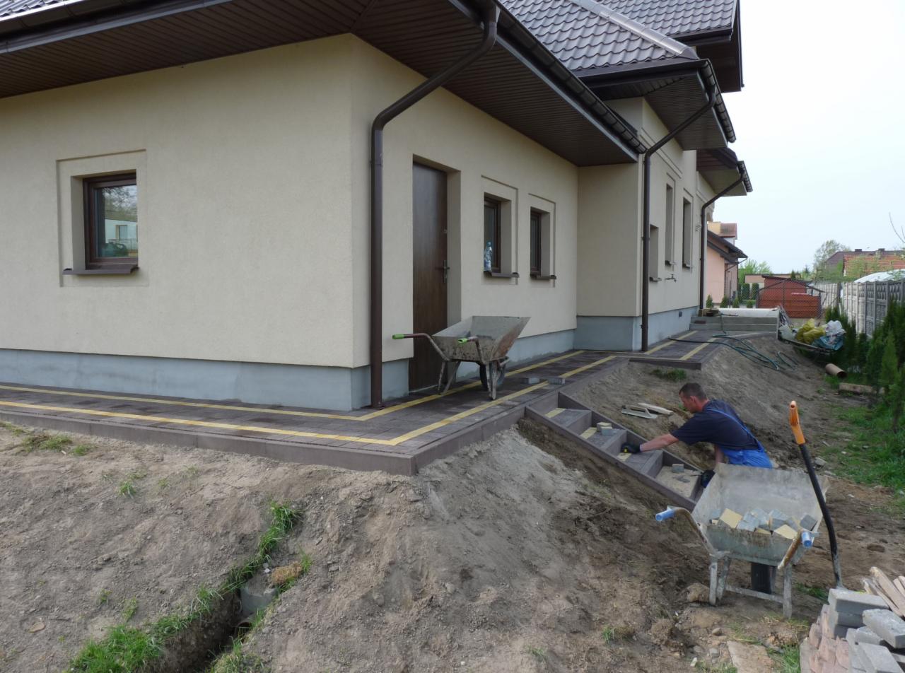 Pracownik w niebieskim ubraniu układa schody z kostki brukowej wzdłuż ściany domu z brązowym dachem, widoczna taczka z kostką i narzędzia.