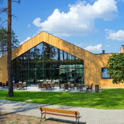 Nowoczesny budynek z drewna o jasnej elewacji z dużymi przeszkleniami, otoczony zielonym trawnikiem, na którym znajdują się stoliki z krzesłami oraz ławka.