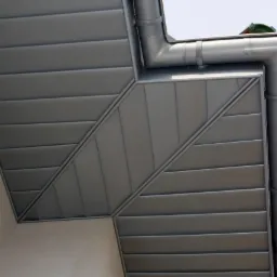 Szara, metalowa rynna narożna, widoczna od dołu, z mocowaniem do ściany budynku. Elementy połączone pod kątem, tworząc estetyczne wykończenie dachu.