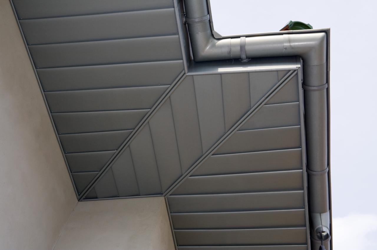 Szara, metalowa rynna narożna, widoczna od dołu, z mocowaniem do ściany budynku. Elementy połączone pod kątem, tworząc estetyczne wykończenie dachu.