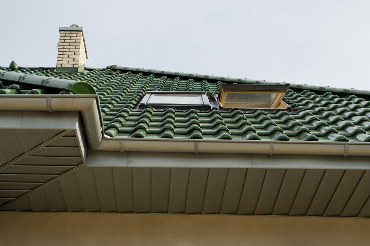 Zielony dach z dachówką karpiówką, otwartym oknem dachowym i szarą rynną, zwieńczony ceglanym kominem na tle jasnego nieba.