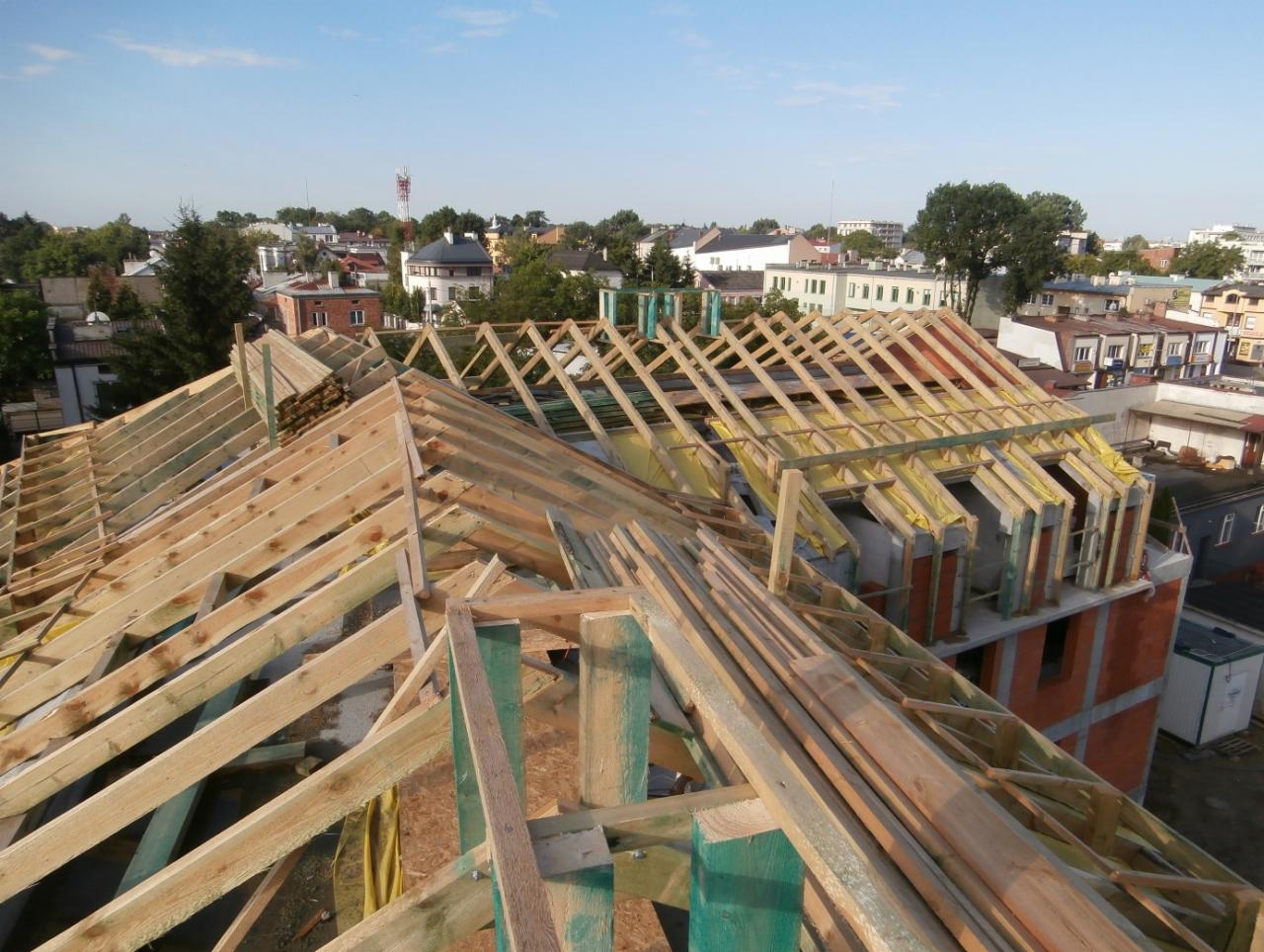 Wznoszenie drewnianej więźby dachowej na tle zabudowy miejskiej, widok z góry. Elementy konstrukcyjne zabezpieczone żółtą folią. Etap budowy dachu.
