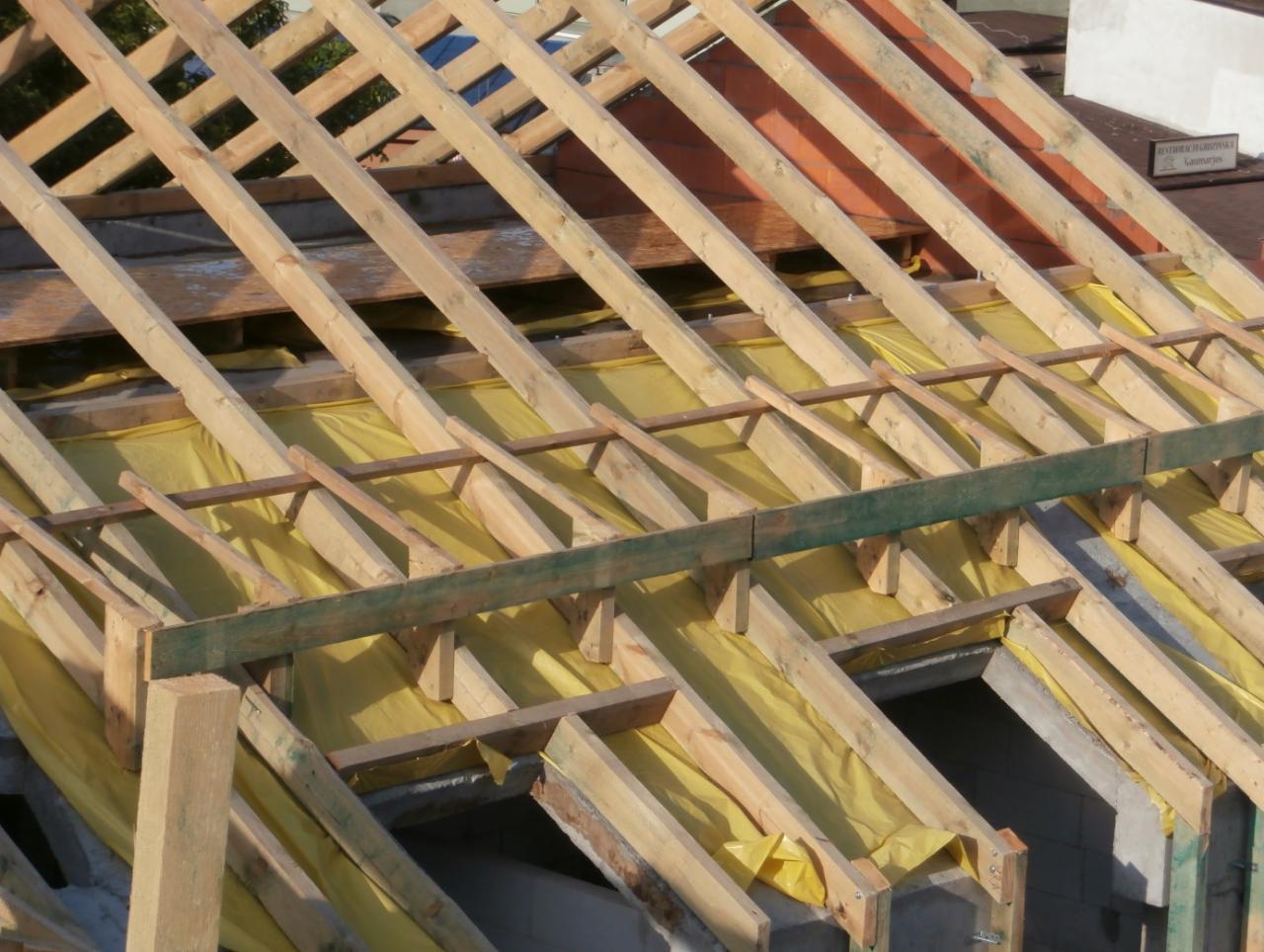 Wiązar dachowy z żółtą membraną w trakcie budowy. Widoczne drewniane elementy konstrukcyjne i fragment budynku w tle. Ujęcie z góry, w dziennym świetle.