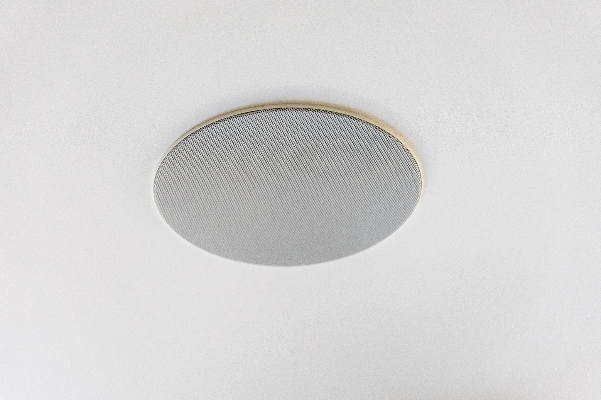 Okrągły głośnik sufitowy z metalową siatką ochronną, umieszczony na białym suficie. Widoczny z bliska, z delikatnym cieniem podkreślającym jego kształt.