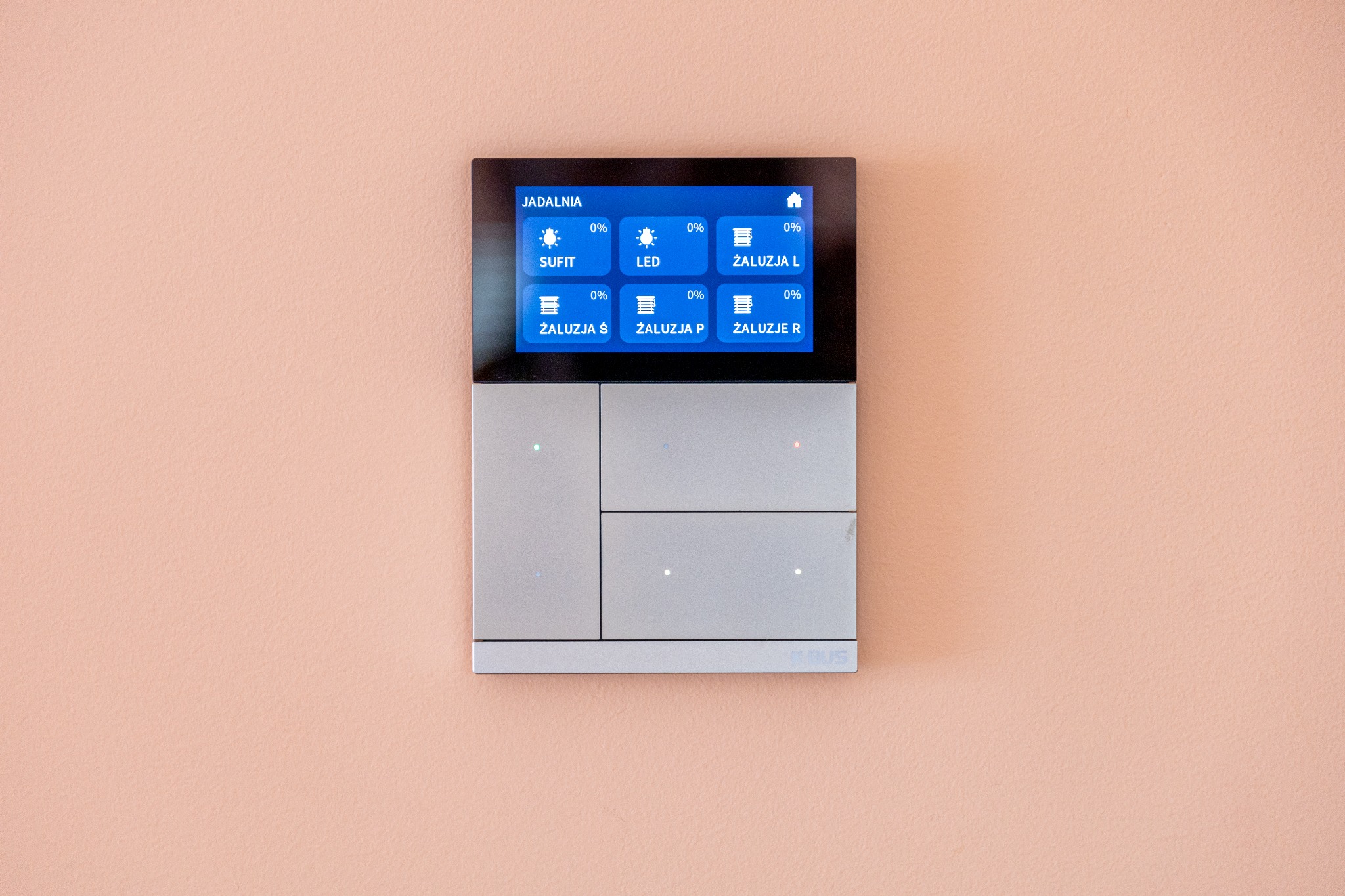 Panel sterowania inteligentnego domu na ścianie w kolorze brzoskwini, z dotykowym ekranem pokazującym kontrolę oświetlenia (Sufit, LED) i rolet (Żaluzja L, Żaluzja Ś, Żaluzja P, Żaluzje R) w jadalni.