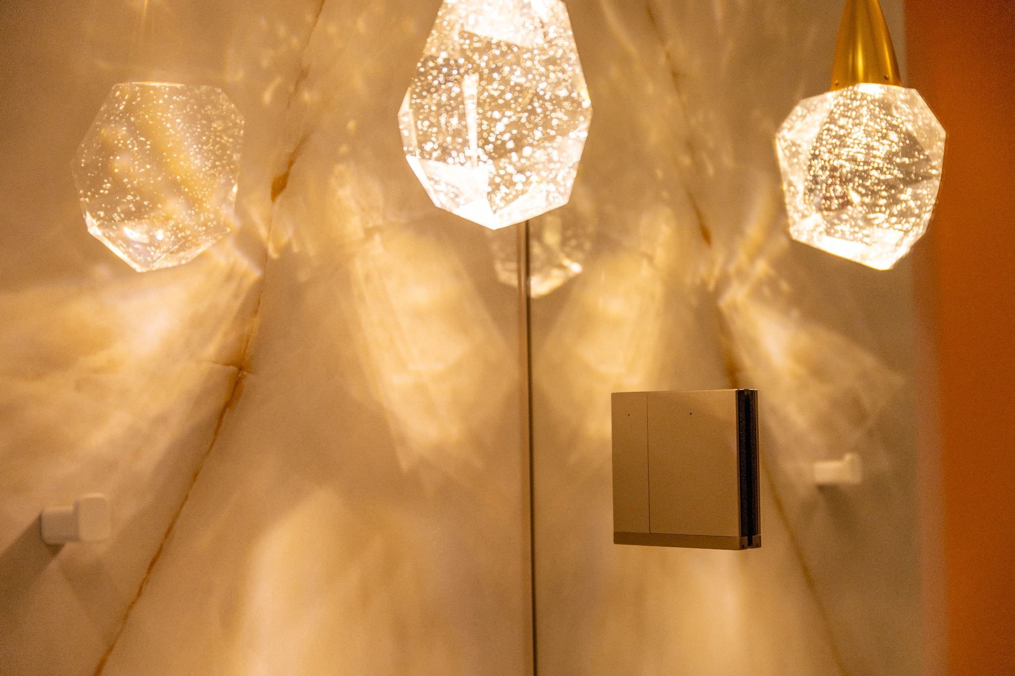 Trzy kryształowe lampy wiszące z ciepłym światłem odbijającym się na marmurowej ścianie, obok nowoczesny panel sterowania oświetleniem z subtelnymi czerwonymi diodami.