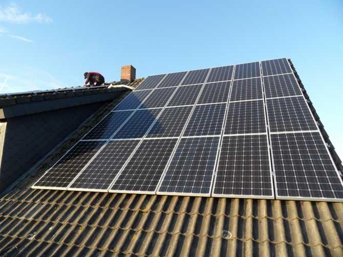 Instalacja paneli słonecznych na dachu domu z ciemnej dachówki; monter w czerwonej kurtce pracuje przy kominie.