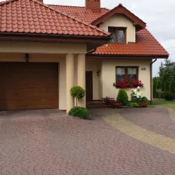 Dom jednorodzinny z czerwonym dachem i garażem, beżowa elewacja, zadbany ogród z tujami i kostką brukową, widoczny samochód osobowy.