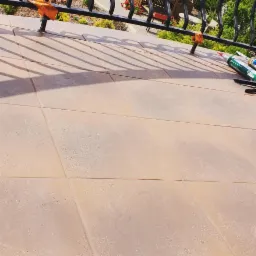 Układanie beżowych płytek na balkonie, widoczne pomarańczowe kliny dystansowe, czarna balustrada i tuby z klejem.