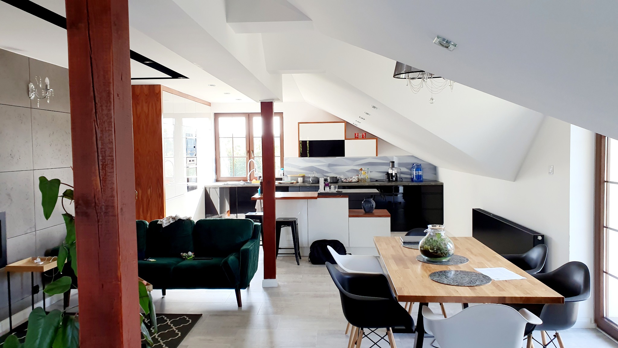 Jasne wnętrze mieszkania na poddaszu z drewnianą kolumną, zieloną sofą, czarno-białą kuchnią i drewnianym stołem jadalnym z czarnymi i białymi krzesłami.