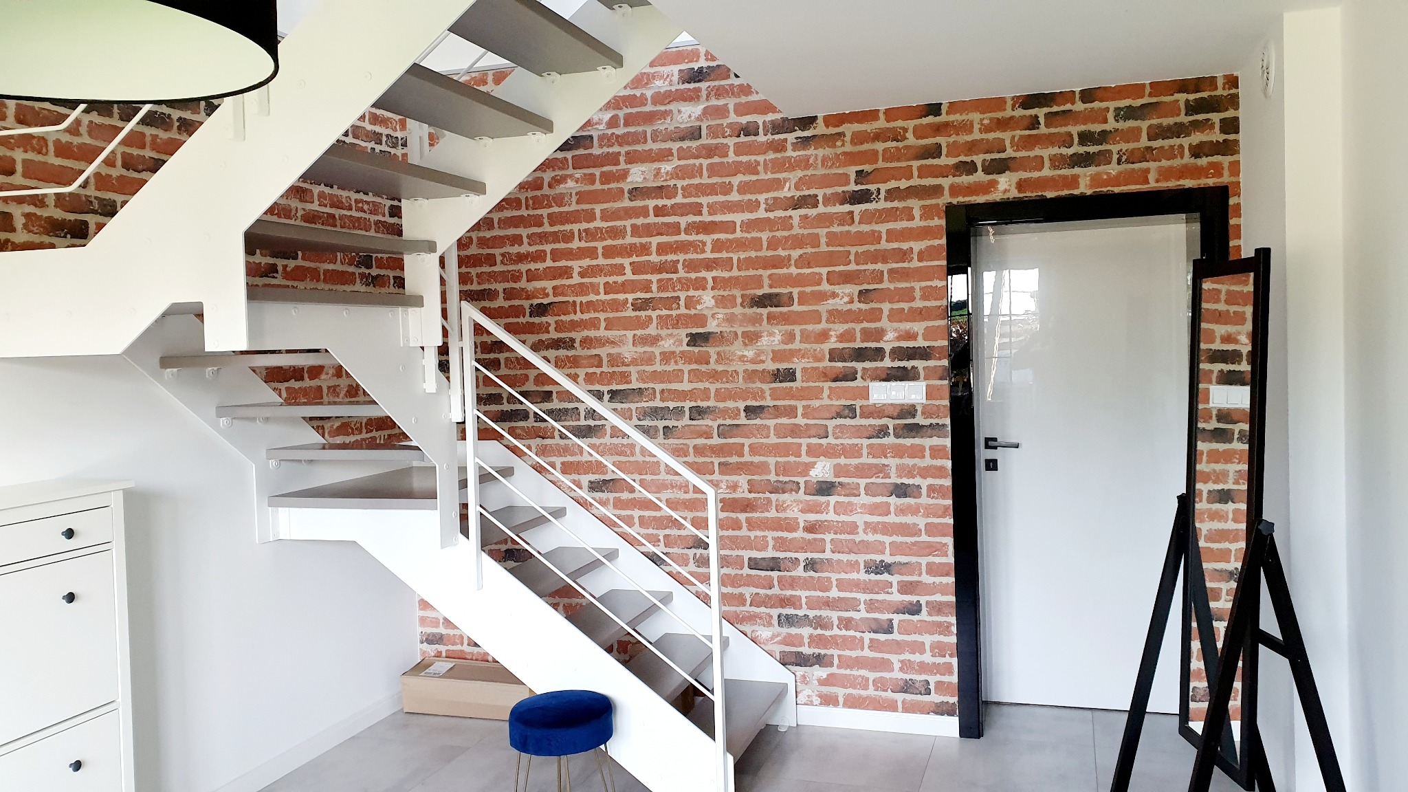 Nowoczesne schody metalowe z drewnianymi stopniami, białe ściany, dekoracyjna ściana z cegły, białe drzwi z czarną ramą i minimalistyczne lustro w przedpokoju.