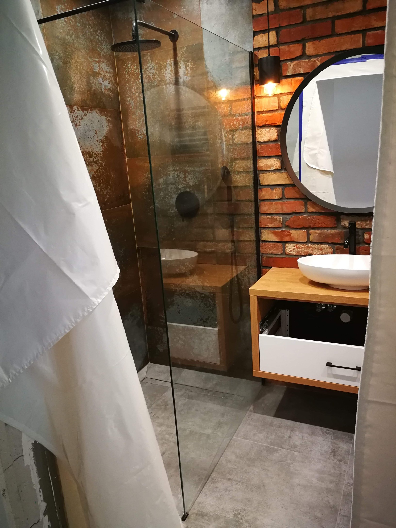 Nowoczesna łazienka z kabiną prysznicową typu walk-in, umywalką nablatową na drewnianej szafce z otwartą szufladą i lustrem w okrągłej, czarnej ramie na tle ściany z czerwonej cegły.