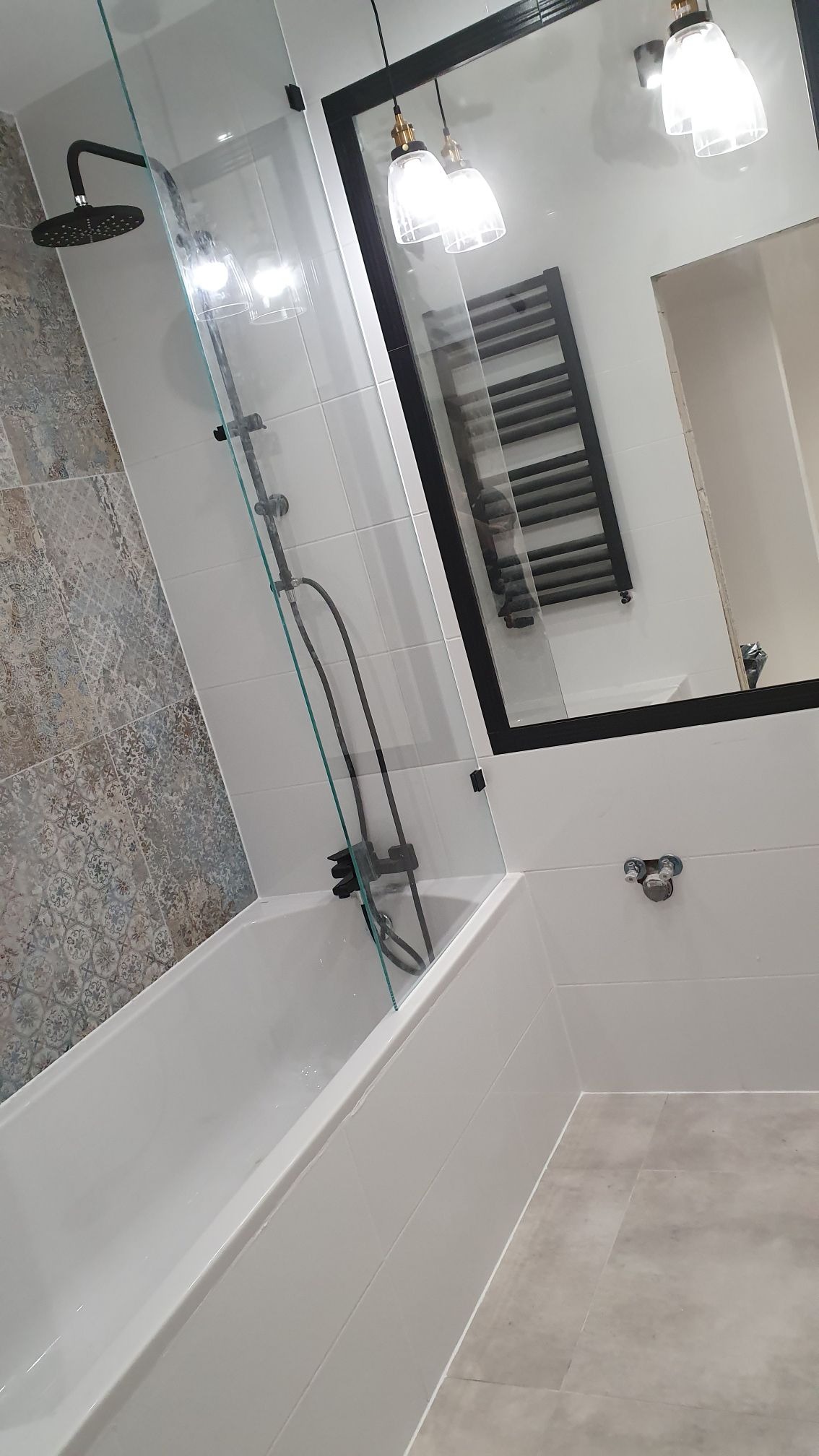 Nowoczesna łazienka po remoncie z białą wanną, szklanym parawanem, czarną armaturą i grzejnikiem drabinkowym, odbijająca się w lustrze podwieszanym pod sufitem.