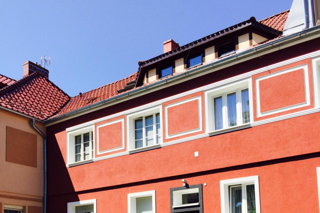 Czerwona fasada budynku z białymi obramowaniami okien i dekoracyjnymi panelami. Widoczny fragment dachu z czerwonej dachówki i błękitne niebo.