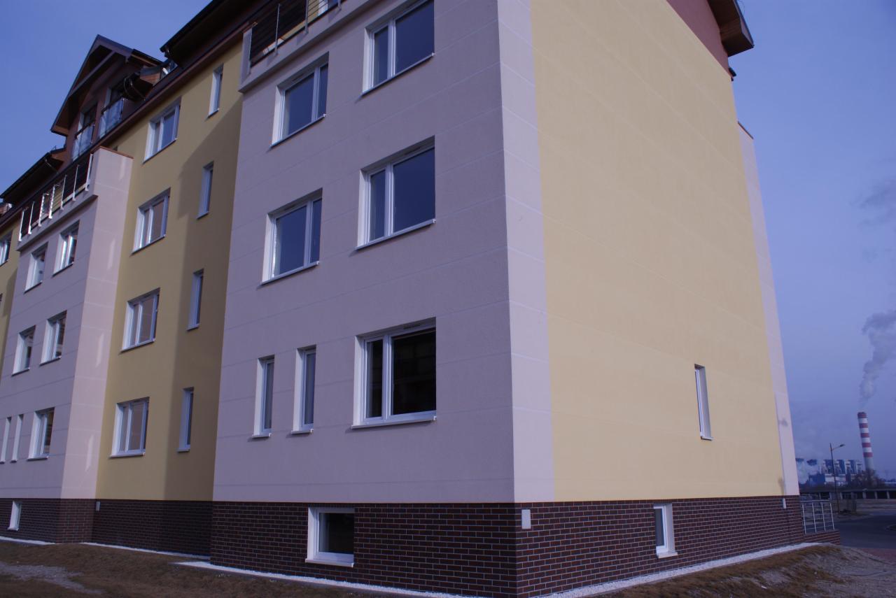 Nowoczesny budynek wielorodzinny po renowacji elewacji. Widoczna dwukolorowa fasada, okna PCV i cokół z cegły klinkierowej. W tle komin przemysłowy.