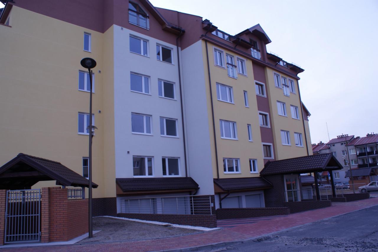 Wielopiętrowy budynek mieszkalny z elewacją w odcieniach żółci, bieli i brązu, widoczny z zewnątrz, z lampą uliczną i fragmentem chodnika z czerwonej kostki.