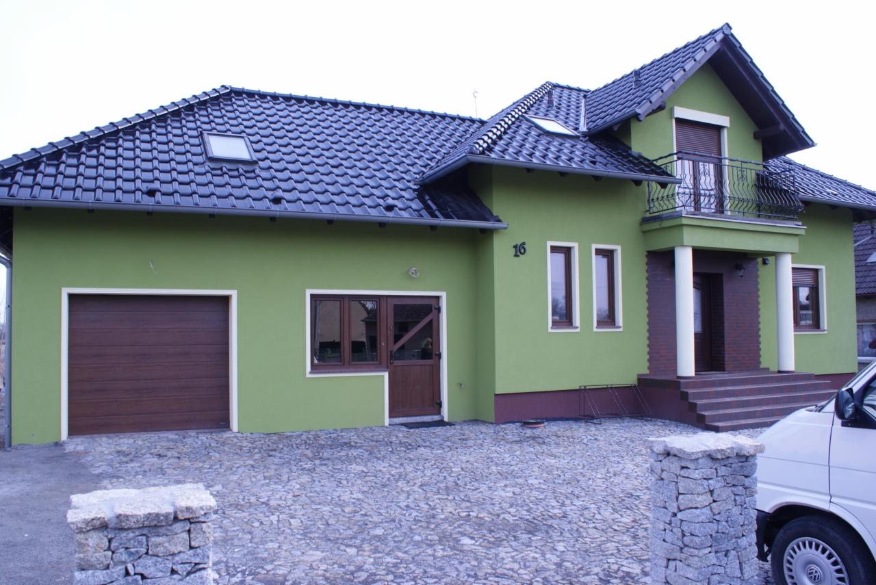 Dwukondygnacyjny dom jednorodzinny z zieloną elewacją, ciemną dachówką i balkonem z kutą balustradą, widoczny z poziomu brukowanego podjazdu.