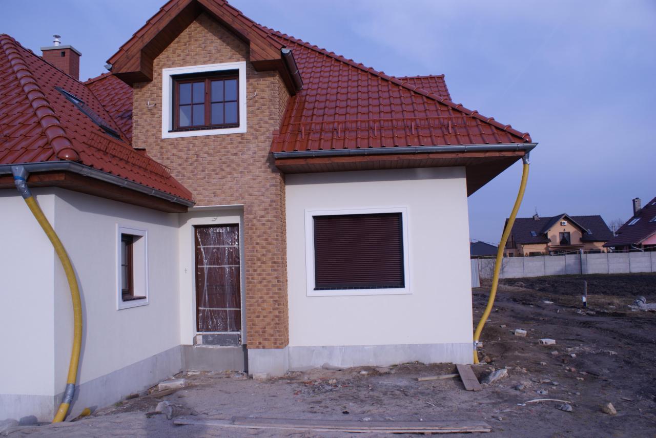 Nowo wybudowany dom jednorodzinny z czerwonym dachem, elewacją z jasnego tynku i fragmentami z cegły, zabezpieczone drzwi wejściowe folią, widoczne rury spustowe.