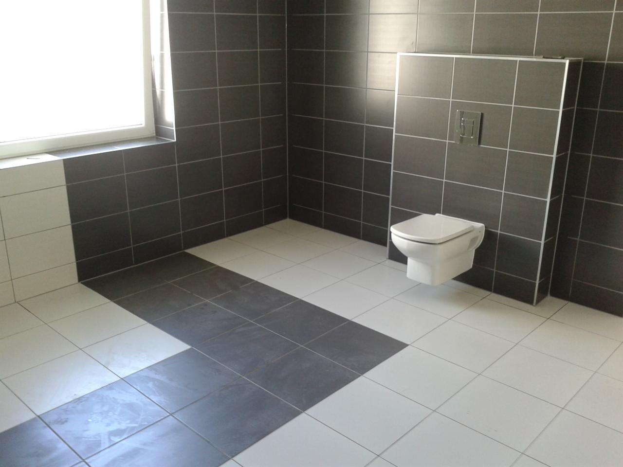 Wykończona łazienka z szarymi płytkami na ścianach i podwieszaną toaletą, białe płytki na podłodze z szarym pasem, okno z lewej strony.