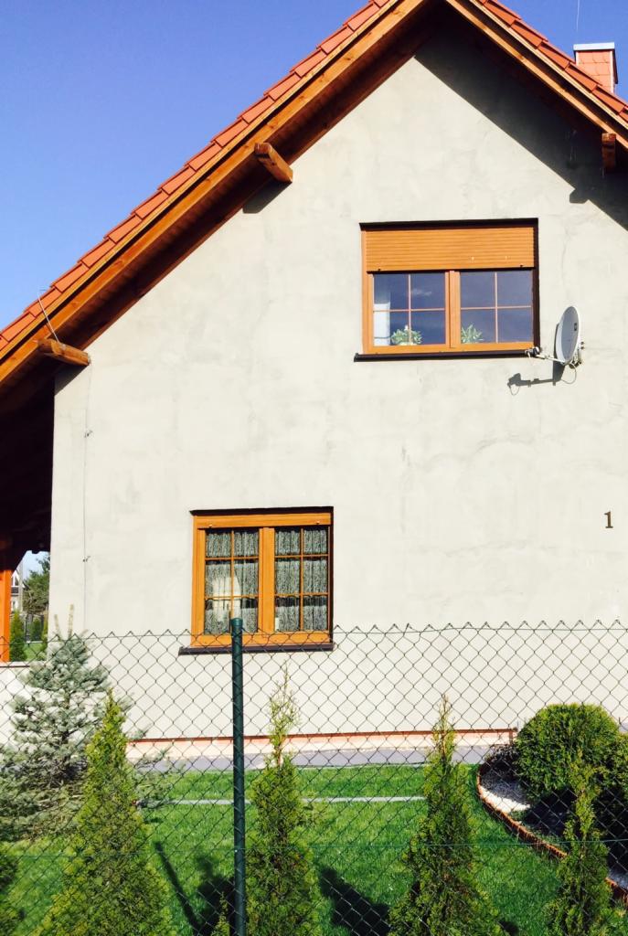 Fragment elewacji domu jednorodzinnego z brązową dachówką, drewnianymi oknami, anteną satelitarną i ogrodzeniem z siatki.