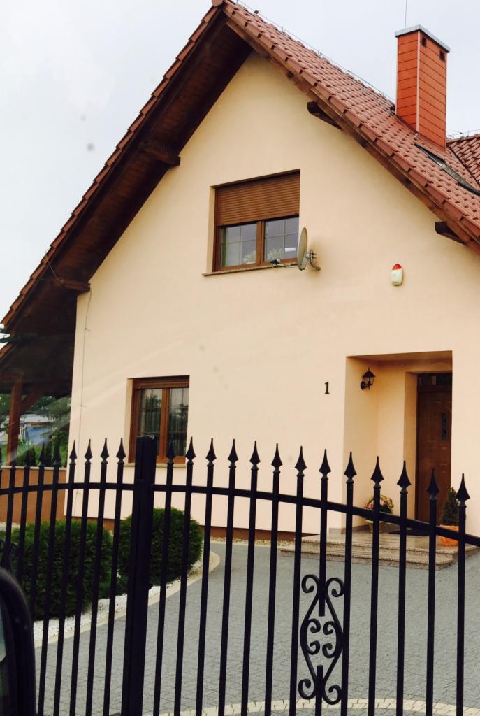 Jasna elewacja domu jednorodzinnego z brązowymi oknami i roletą, widoczna zza czarnego, kutego ogrodzenia z ozdobnymi elementami.