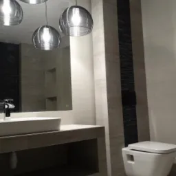 Nowoczesna łazienka w odcieniach szarości z wiszącą toaletą, umywalką nablatową i trzema metalowymi lampami wiszącymi o ażurowej konstrukcji.