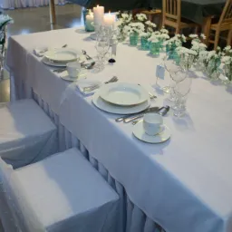 Uroczysta dekoracja stołu z białym obrusem, pokrowcami na krzesła z siateczki w kropki, białymi kwiatami w małych wazonach i świecami w szklanych lampionach.