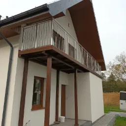 Nowoczesny balkon z prostą, stalową balustradą w Kołobrzegu. Widoczne drewniane elementy konstrukcyjne balkonu i fragment elewacji budynku. Na dole, przy ścianie, stoją puste pojemniki po...